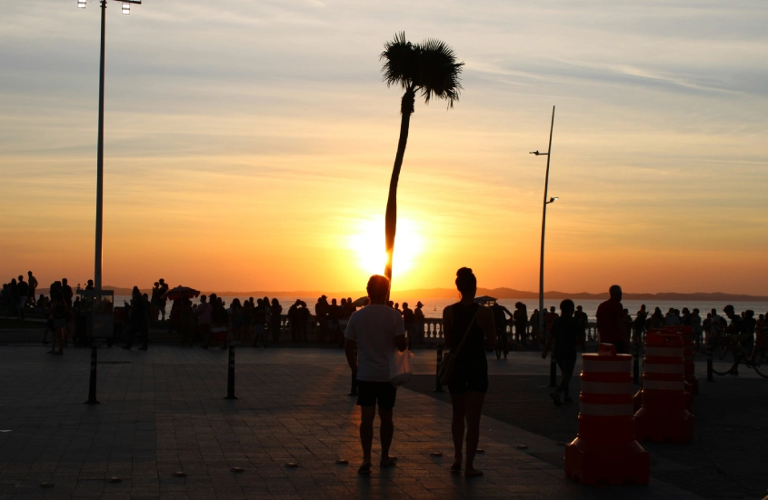 Pôr do sol na orla de Salvador com silhuetas de pessoas, uma palmeira destacada no centro e o céu em tons de laranja e dourado.