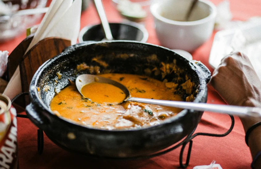 Panela de moqueca baiana em uma mesa com toalha vermelha, destacando o caldo alaranjado com ervas e uma concha prateada.