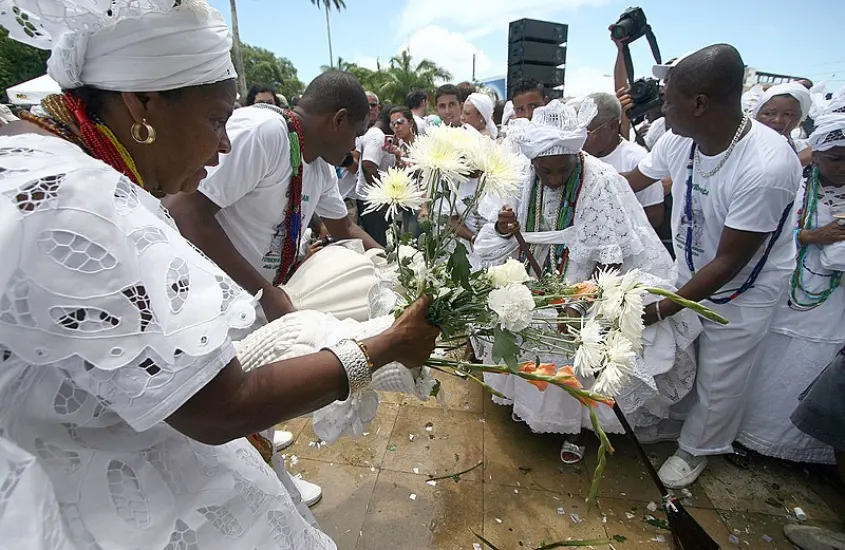 Celebração religiosa em Salvador, com participantes vestidos de branco segurando flores em homenagem à Iemanjá.