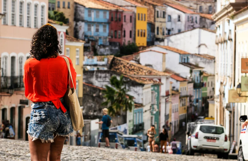 Mulher de blusa vermelha e short jeans caminhando pelo Pelourinho, com casarios coloridos ao fundo em Salvador.