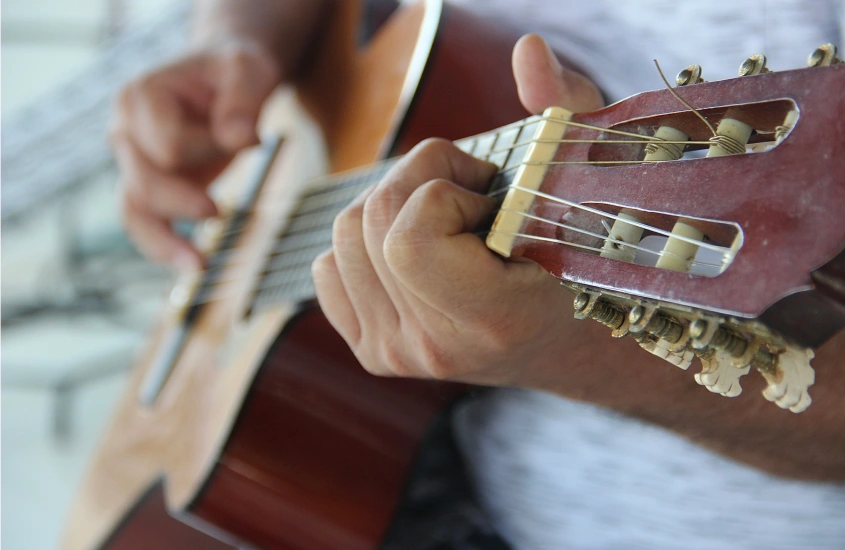 Mãos tocando um violão de madeira em um ambiente descontraído, com foco nas cordas e no movimento dos dedos.