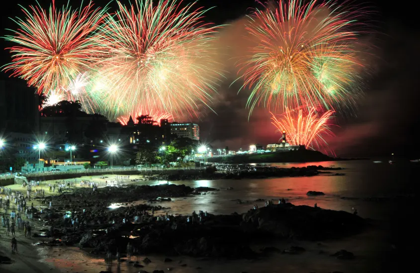 Fogos de artifício iluminando a noite na Praia do Farol da Barra, em Salvador, com reflexos coloridos no mar.