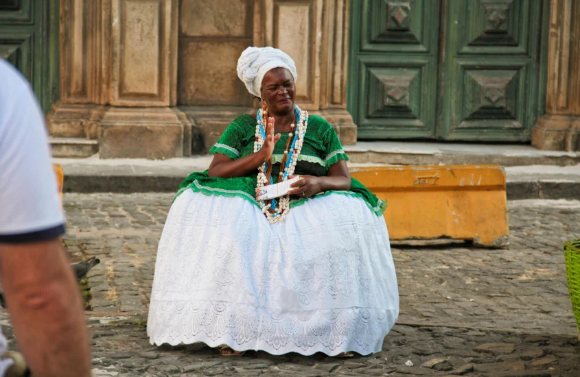 Mulher vestida com traje tradicional baiano, em tons de verde e branco, sentada em frente a uma porta de madeira histórica.