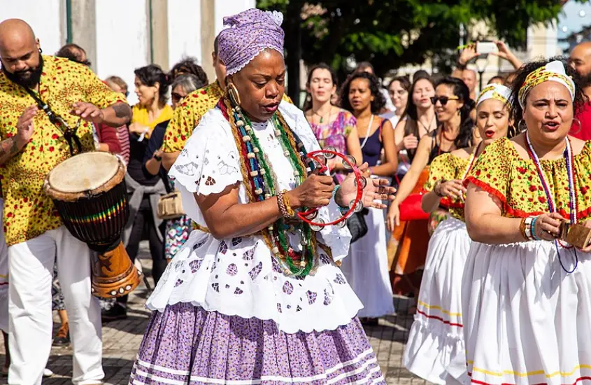 Grupo cultural baiano performando com tambor e dança tradicional, com figurinos coloridos e público ao fundo.