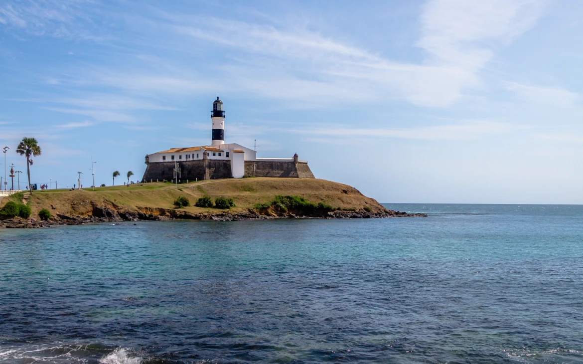 Farol da Barra em Salvador, erguido sobre um pequeno morro gramado, cercado pelo mar azul e céu limpo ao fundo.