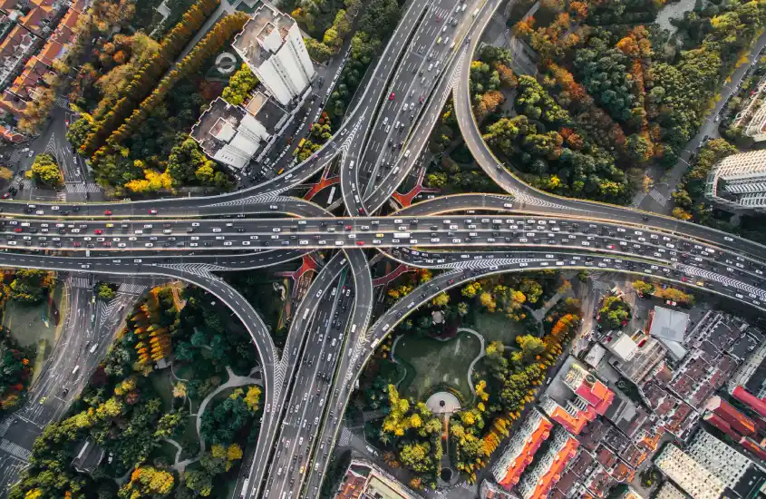 Vista aérea de um complexo de rodovias entrelaçadas, com carros em movimento e rodeadas por áreas verdes e prédios altos.