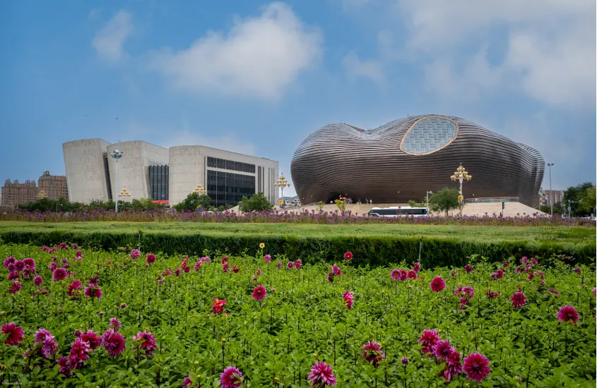 Estrutura moderna e arrojada de um prédio em forma de concha ao lado de um edifício de design quadrado, com um jardim florido em primeiro plano e céu azul claro.