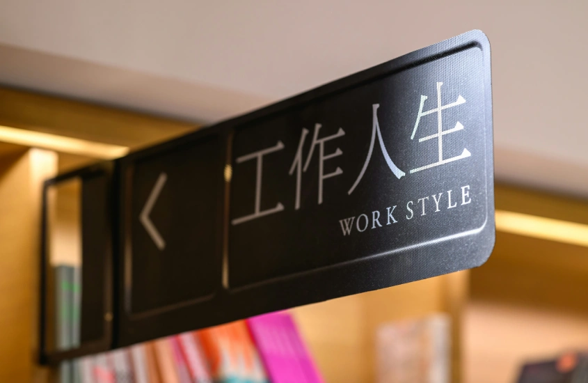 Placa de sinalização com caracteres chineses e a inscrição “Work Style” em inglês, com prateleiras e livros desfocados ao fundo.