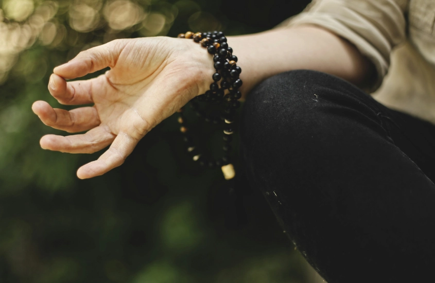 Mão de uma pessoa em posição meditativa, com um rosário enrolado no pulso, contra um fundo verde desfocado, sugerindo um momento de paz e concentração.