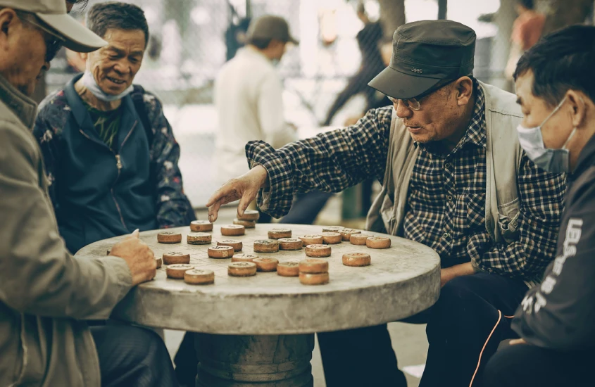 Grupo de homens idosos jogando um jogo de tabuleiro tradicional em uma mesa de pedra ao ar livre, em um ambiente descontraído.