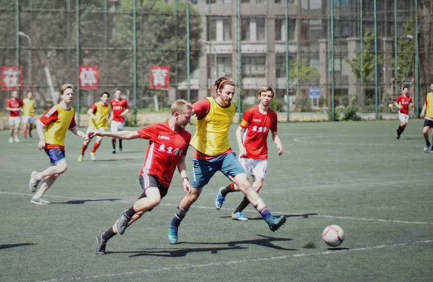 Jogo de futebol em andamento em um campo de grama sintética, com jogadores usando uniformes coloridos e disputando a posse da bola.