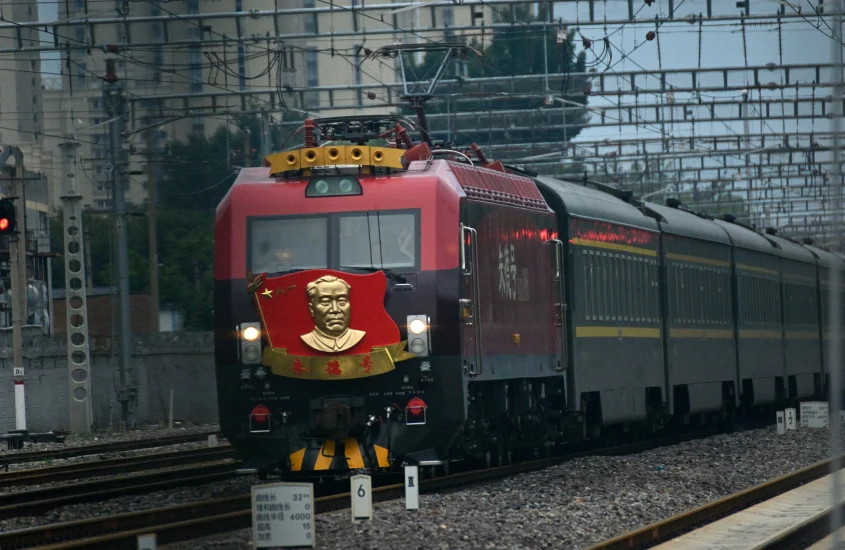 Trem vermelho com um emblema de Mao Tse Tung na frente, em uma ferrovia, em movimento sob a estrutura de fios e postes elétricos.