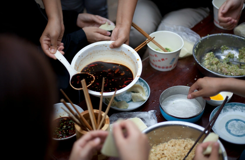 Pessoas ao redor de uma tigela grande com molho, utilizando hashi e preparando dumplings em um ambiente familiar.