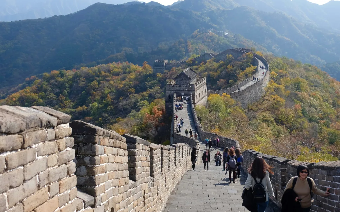 Turistas caminhando ao longo de um trecho da Grande Muralha da China, cercados por paisagens montanhosas e vegetação colorida de outono