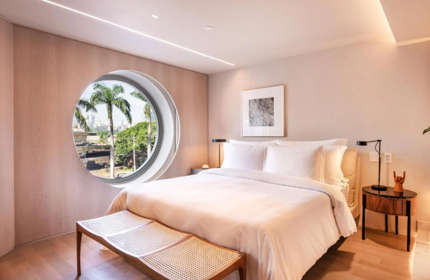 Quarto moderno com cama de casal e janela circular que enquadra palmeiras e uma vista urbana ao fundo.