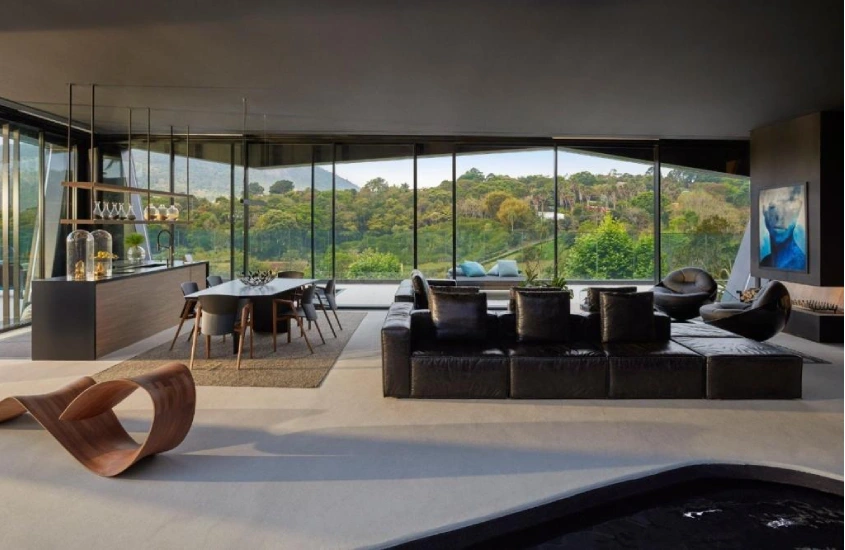 Sala de estar contemporânea com sofás de couro preto, mesa de jantar e grandes janelas de vidro que oferecem uma vista para a vegetação ao redor.