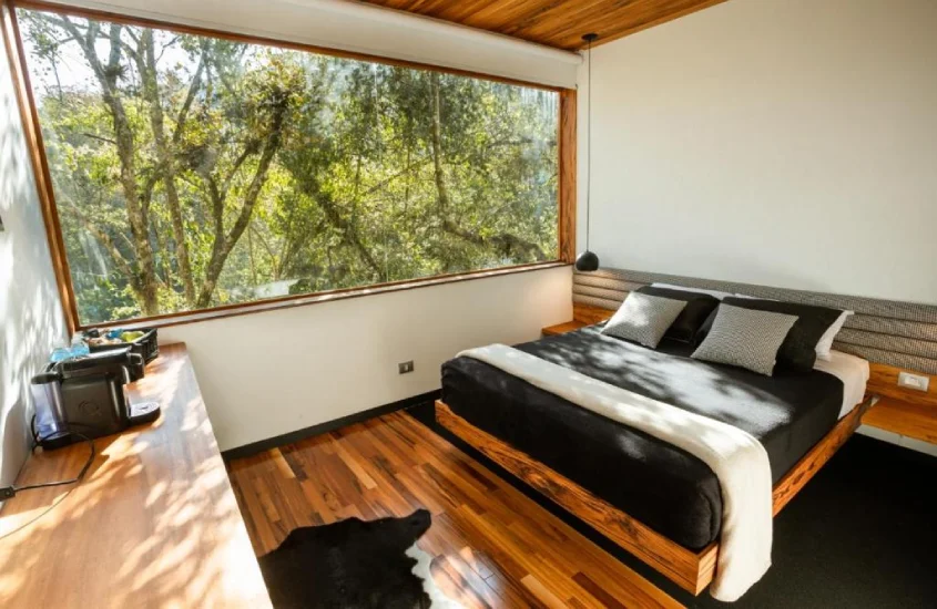 Quarto aconchegante com cama de casal e detalhes em madeira, com uma janela ampla que exibe a natureza ao redor.