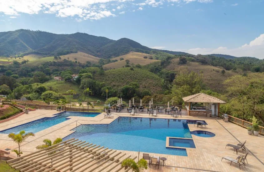 Piscina ao ar livre com espreguiçadeiras e mesas, cercada por colinas e vegetação exuberante, com montanhas ao fundo.