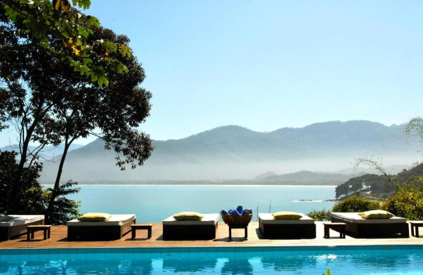 Piscina ao ar livre com espreguiçadeiras, rodeada por árvores e uma vista incrível para o mar e montanhas ao fundo.