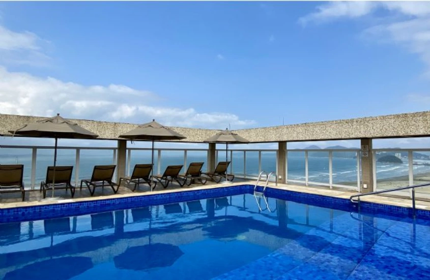 Piscina no rooftop com espreguiçadeiras e guarda-sóis ao redor, oferecendo uma vista do oceano em um cenário ensolarado.