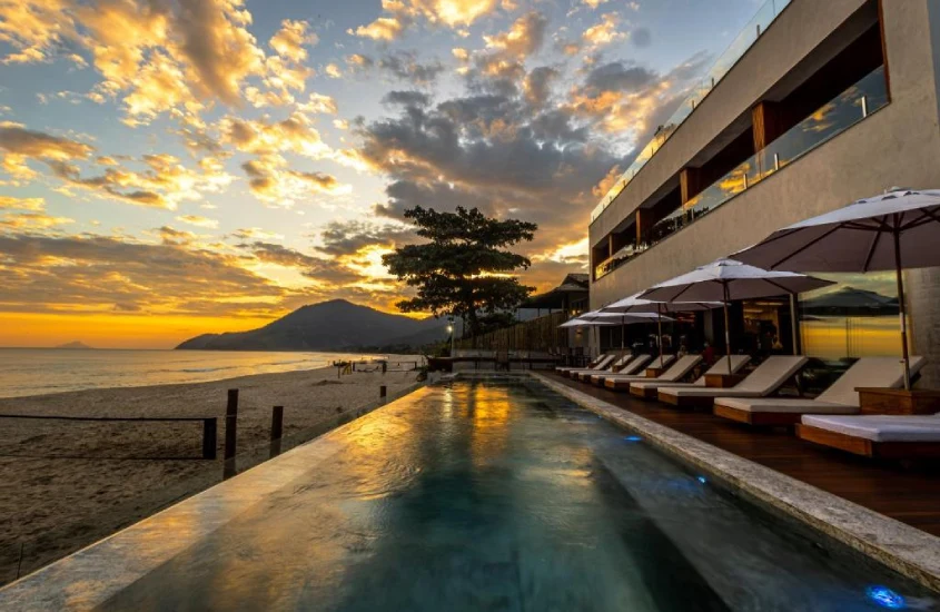 Piscina de borda infinita ao pôr do sol, com espreguiçadeiras e uma vista deslumbrante para o mar e as montanhas ao fundo.