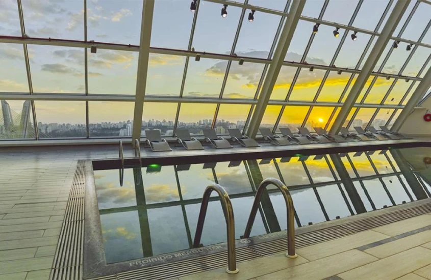 Piscina de borda infinita coberta com espreguiçadeiras ao redor e uma ampla vista da cidade ao entardecer.