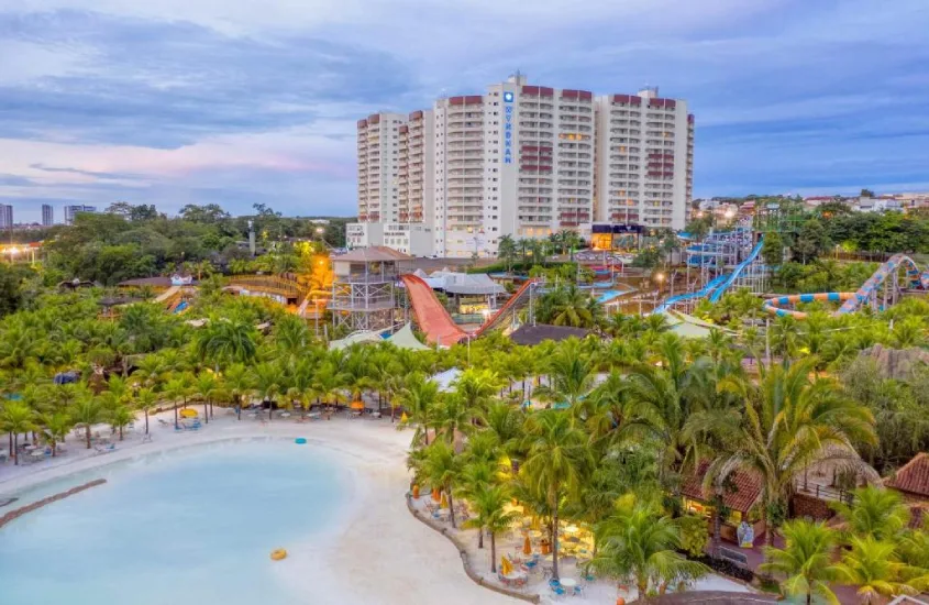 Parque aquático com várias atrações e toboáguas, cercado por vegetação tropical, com um grande prédio ao fundo.