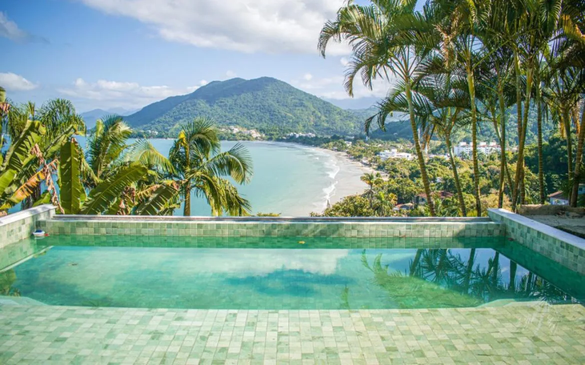 Piscina de borda infinita cercada por palmeiras, com uma vista para a praia e as montanhas ao fundo, em um cenário tropical e ensolarado