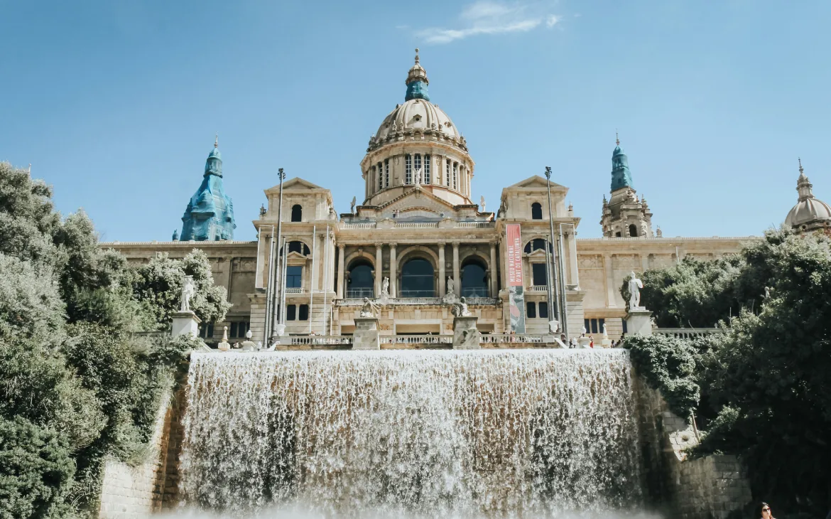 Em um dia de sol, fachada de museu com fonte na frente e árvores ao redor