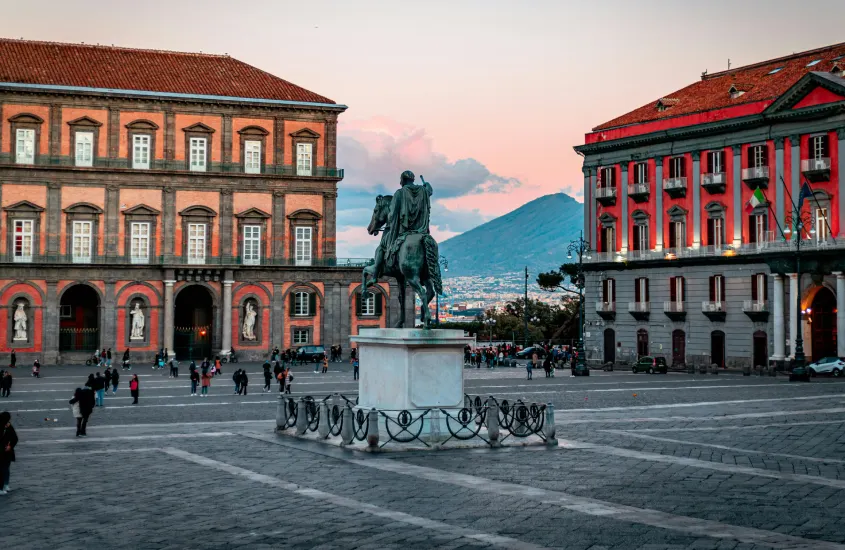 Praça do Plebiscito ao entardecer, com estátua de cavaleiro em primeiro plano e vista do Vesúvio ao fundo, enquanto pessoas passeiam ao redor.