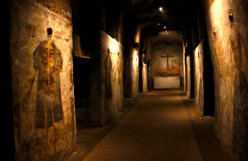 Corredor sombrio de uma catacumba com afrescos de esqueletos em túnicas esculpidos nas paredes de pedra, emoldurados por uma iluminação suave.