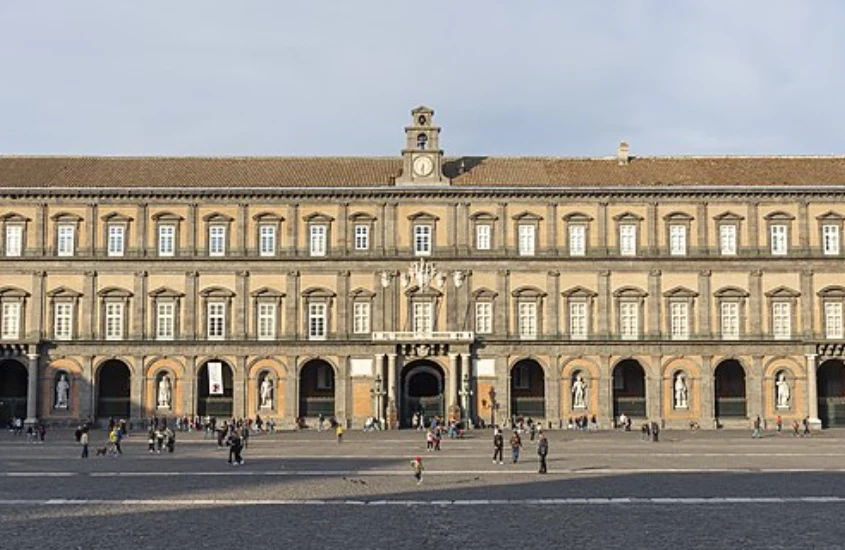 Fachada do Palácio Real de Nápoles em um dia ensolarado, com sua imponente arquitetura simétrica e várias janelas alinhadas.