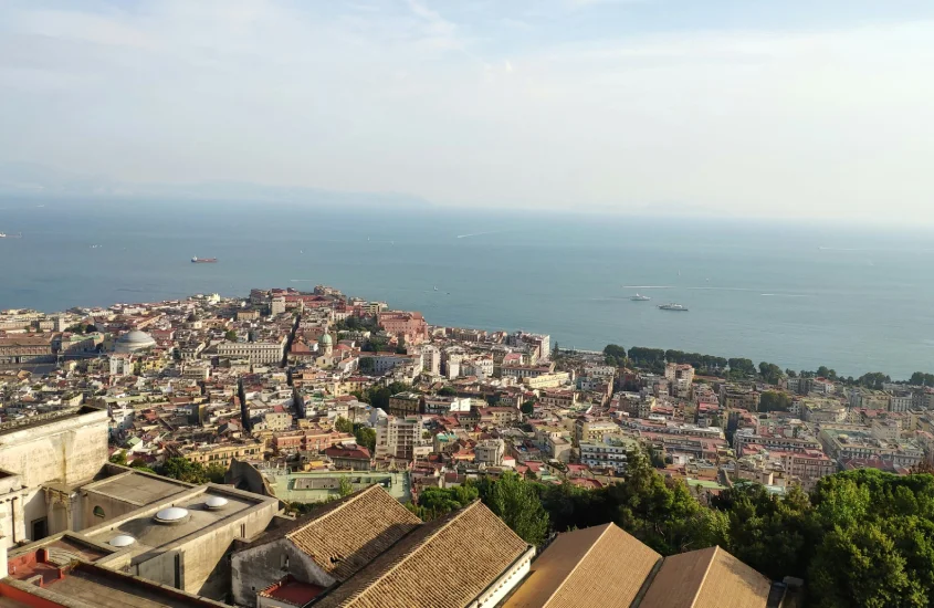 Vista aérea da cidade de Nápoles se estendendo até o mar, com diversas construções e a costa emoldurada pelo céu e o oceano ao fundo.