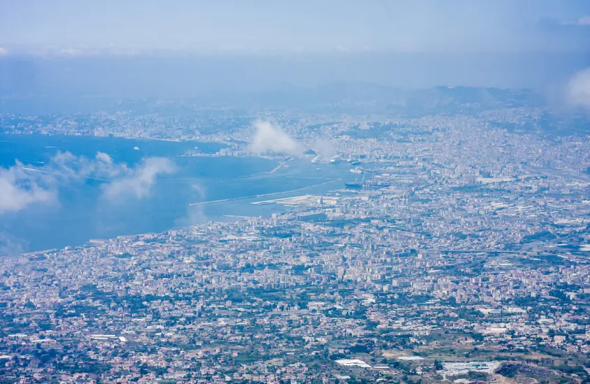 Visão ampla da baía de Nápoles sob um céu azul, com nuvens baixas e a cidade se estendendo ao redor da água.