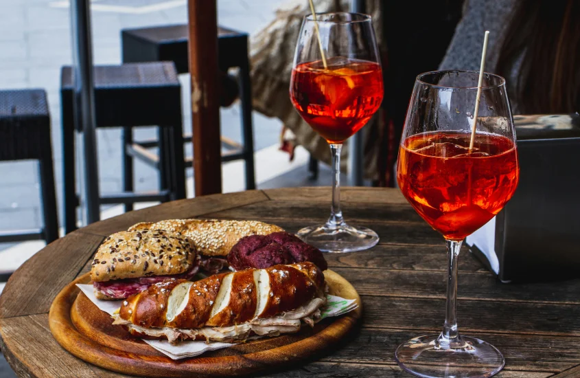 Mesa de madeira com taças de Aperol Spritz e sanduíches artesanais, incluindo pães variados e recheios, em uma típica refeição ao ar livre.