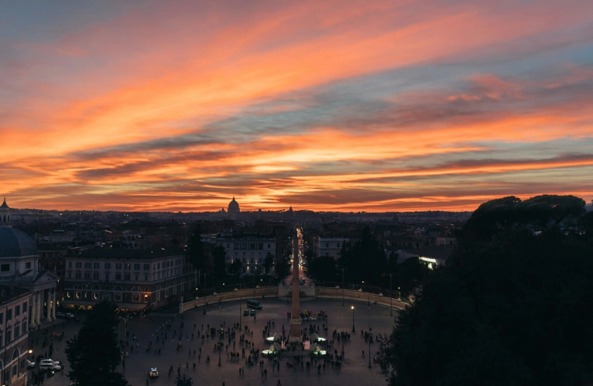 Céu alaranjado ao entardecer sobre a cidade de Roma, com uma vista elevada de uma praça movimentada e a silhueta de edifícios ao fundo.