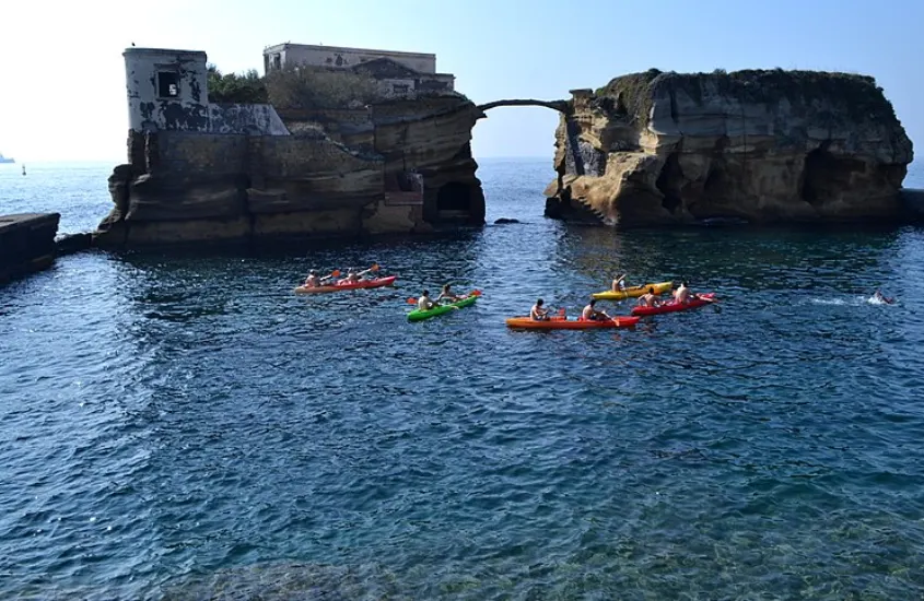 Pessoas remando em caiaques coloridos sobre águas transparentes, explorando formações rochosas e uma ponte de pedra natural sobre o mar.