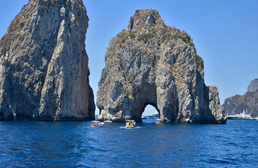 Rochas imponentes dos Faraglioni emergindo do mar azul, com barcos navegando ao redor dessa famosa formação natural em Capri.