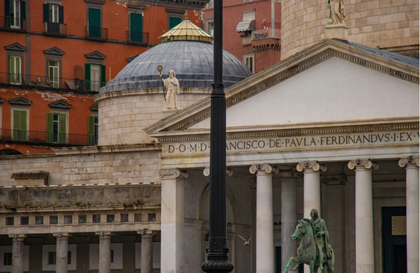 Detalhe da Basílica de San Francesco di Paola, com colunas e cúpula destacando a arquitetura clássica, contra um prédio colorido de Nápoles.