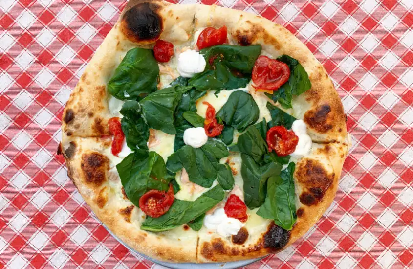 Pizza napolitana com espinafre, tomates e queijo, servida sobre uma toalha de mesa quadriculada em vermelho e branco.