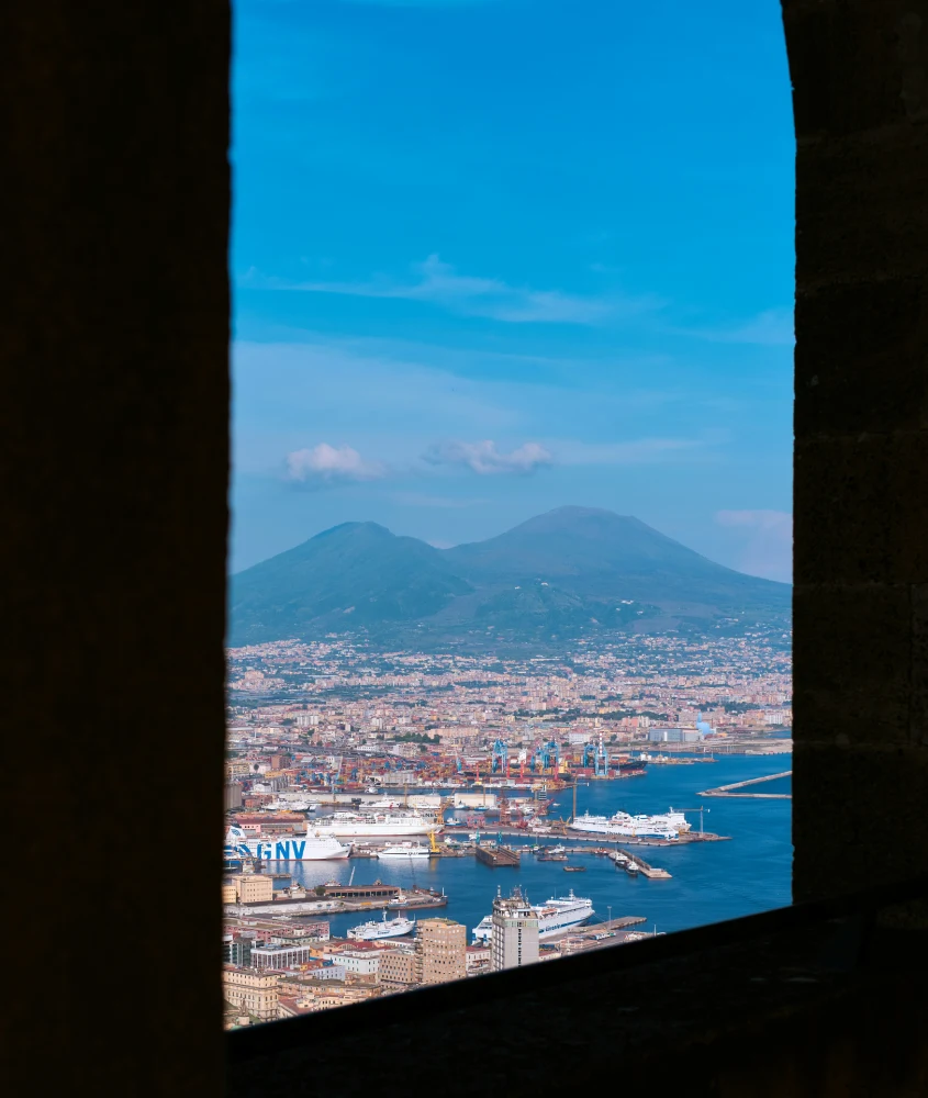 Vista do Golfo de Nápoles e do Vesúvio ao fundo, enquadrada por uma janela de pedra, revelando o porto e parte da cidade.