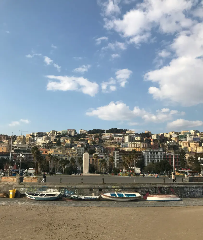 Praia de areia em Nápoles com pequenos barcos ancorados e edifícios residenciais ao fundo em um dia ensolarado.