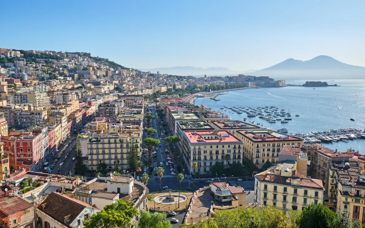 Vista panorâmica de Nápoles em um dia ensolarado, com prédios coloridos ao longo da costa, barcos ancorados no mar e o Monte Vesúvio ao fundo sob um céu azul claro.