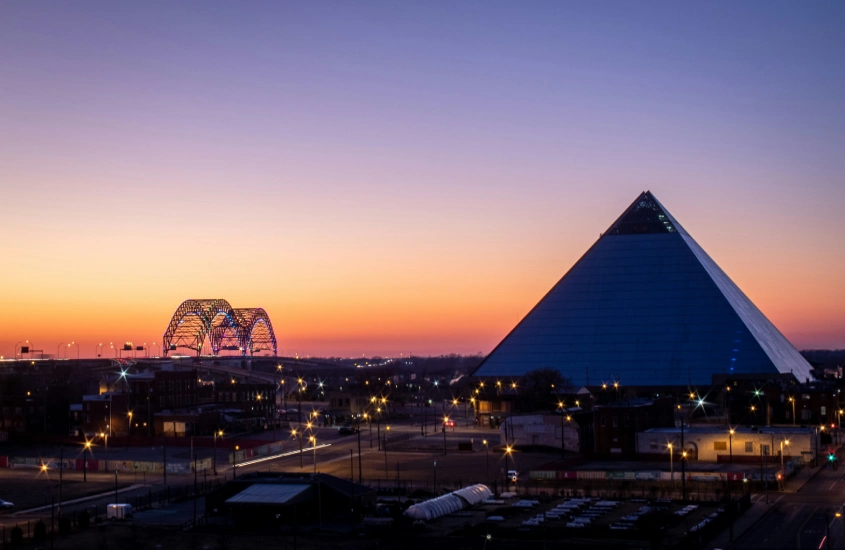 Cena do horizonte de Memphis ao entardecer, com a icônica Pirâmide de Memphis à direita e a ponte Hernando de Soto iluminada ao fundo.