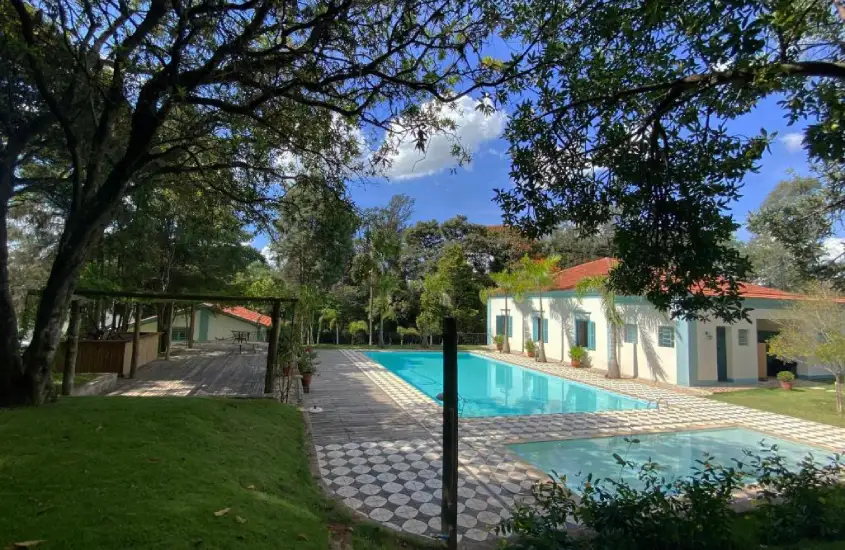 Em um dia de sol, área de lazer de hotel com piscina, casinha do lado e árvores ao redor