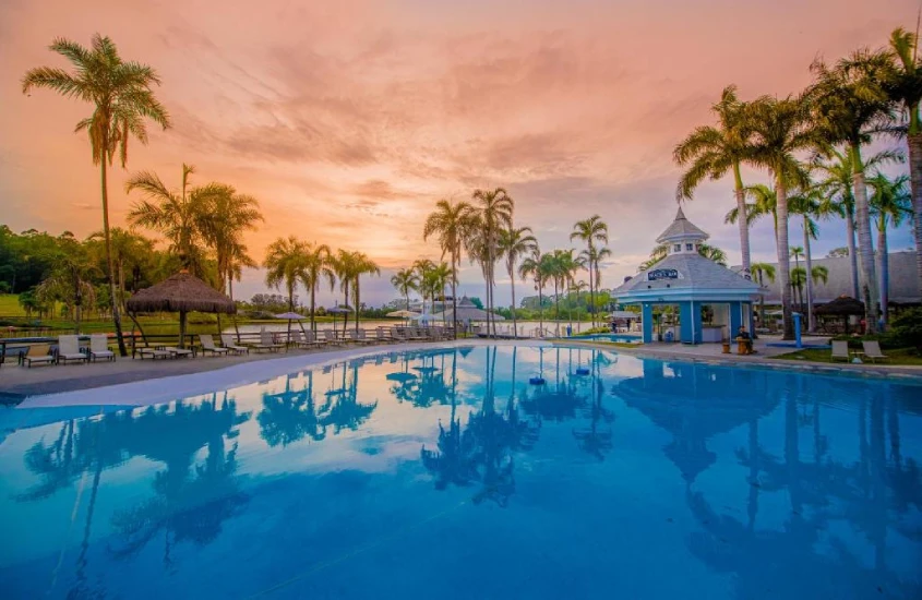 Durante o pôr do sol, área de lazer de um dos hotéis cinco estrelas no interior de SP com piscina, espreguiçadeiras, guarda-sóis coreto e árvores ao redor