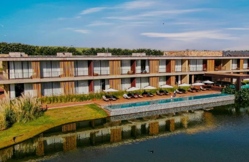 Em um dia ensolarado, vista aérea de um dos hotéis cinco estrelas no interior de SP com lago do lado, piscina, espreguiçadeiras, árvores e plantas decorativas