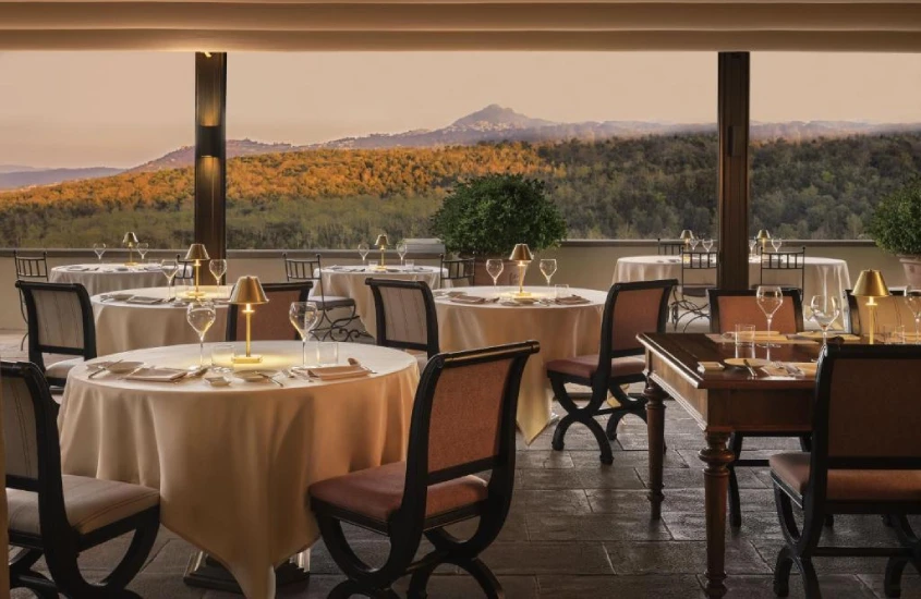 Ambiente elegante com mesas finamente preparadas em um terraço coberto, com vista para as colinas da Toscana iluminadas pela luz do pôr do sol.