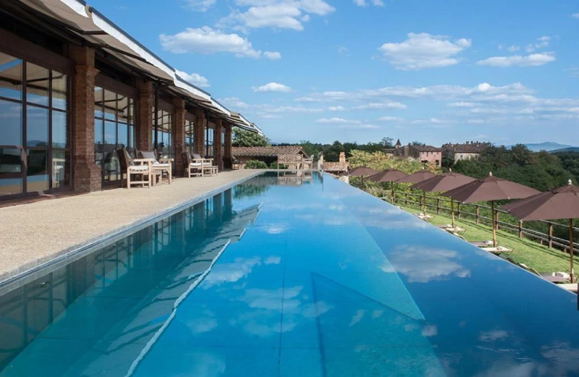 Piscina de borda infinita refletindo o céu azul, com vista para um jardim com guarda-sóis e construções rústicas ao fundo.