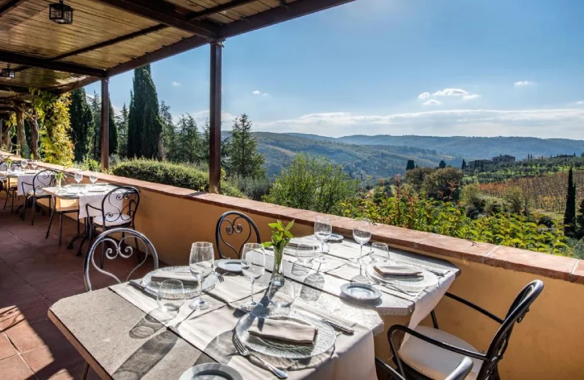 Terraço com mesas elegantemente postas, oferecendo uma vista deslumbrante para os vinhedos e colinas da Toscana em um dia ensolarado.